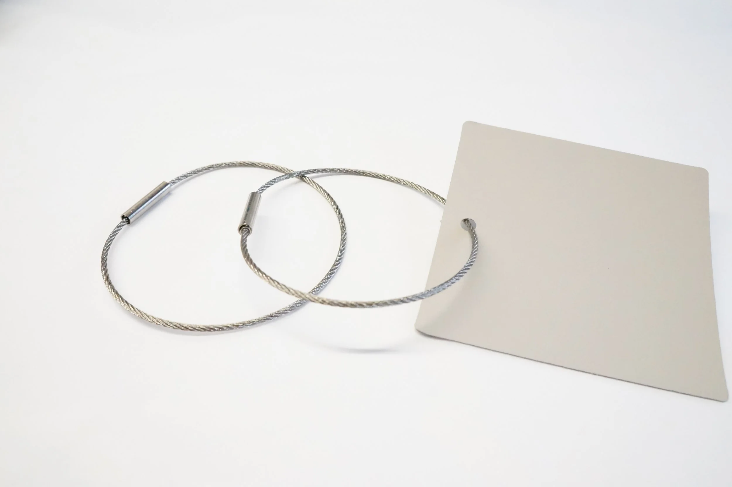 Due braccialetti sottili in metallo con chiusura, posizionati sopra una superficie bianca, uno di essi ha anche un cartoncino bianco attaccato con un foro.