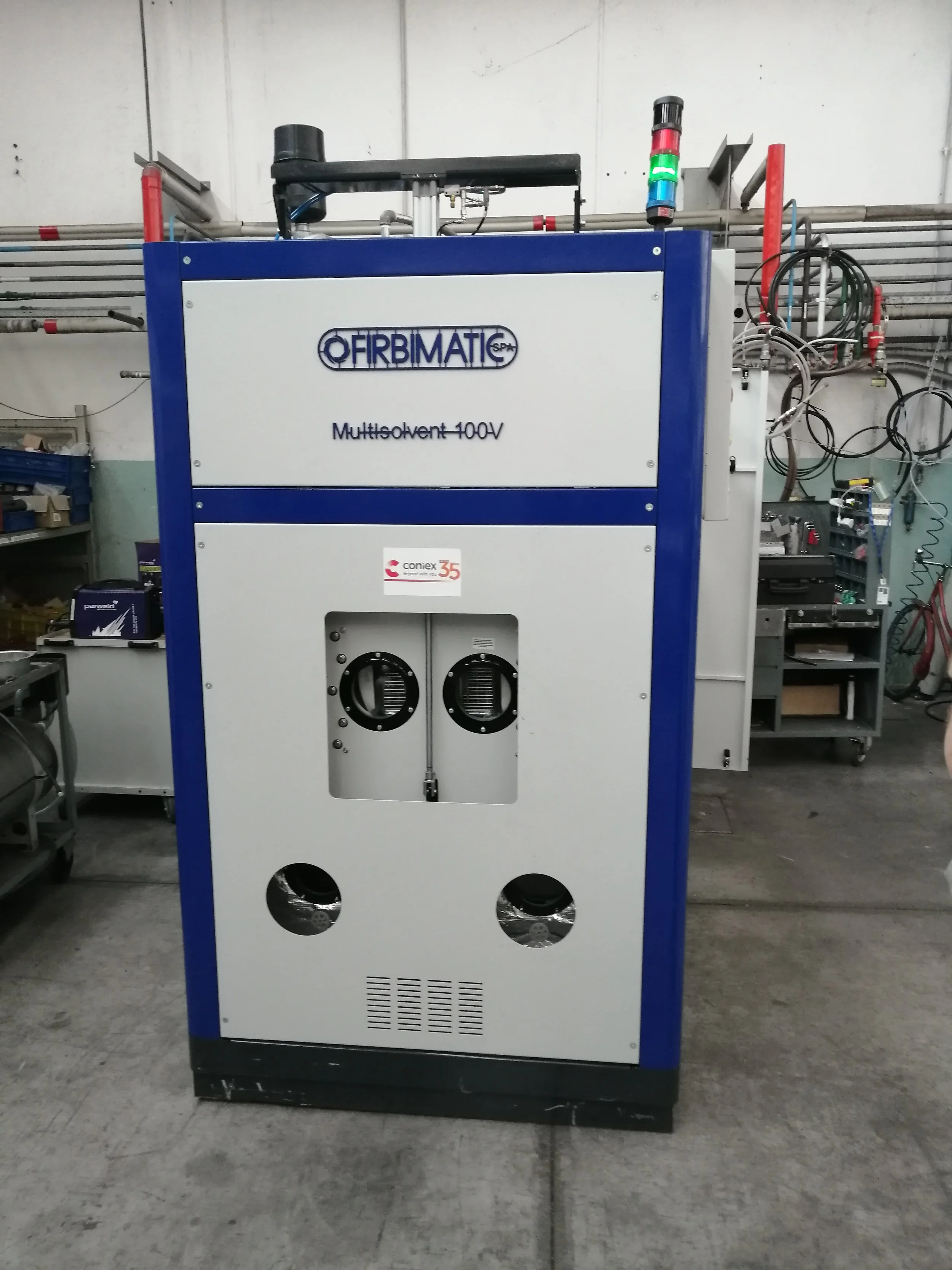 Macchina industriale con etichetta 'FIRBIMATIC' e scritto 'Multisolvent 100V', situata in un ambiente di officina con strumenti e cavi sullo sfondo.